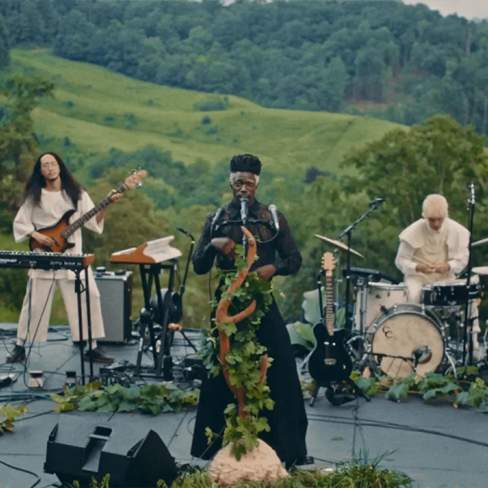 Color photograph of a musical performance outdoors with three musicians playing keyboards, saxophone, and drums against a mountainous green landscape backdrop.