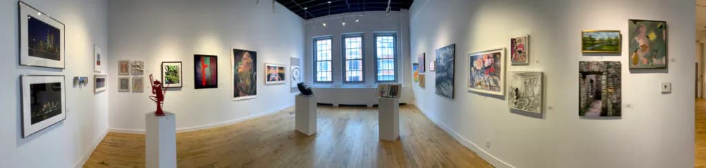 Panoramic view of NOHO M55 Gallery interior with white walls filled with framed artworks, wooden floors, sculpture pedestals, and large windows at the back