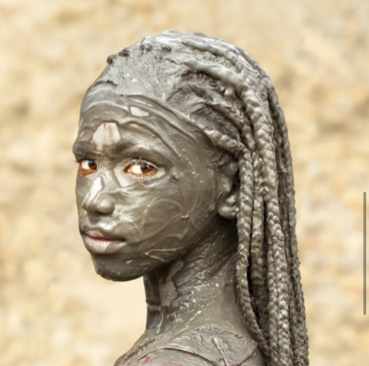 Close-up bronze sculptural portrait of a figure with braided hair and copper-colored eyes against a textured neutral background.