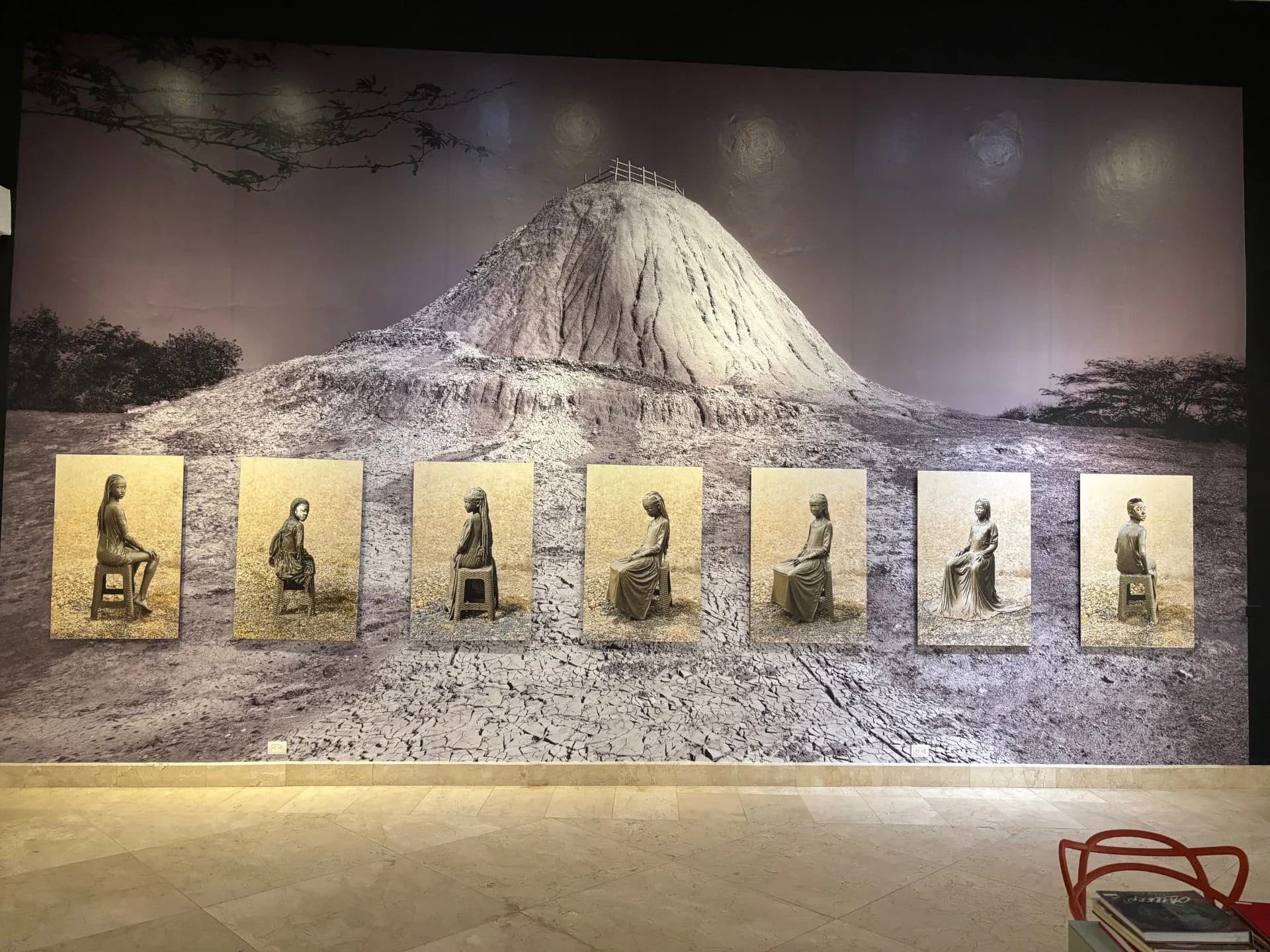 Gallery installation view with a large landscape photograph of a cone-shaped mountain and seven framed seated figure photographs on a cream wall.