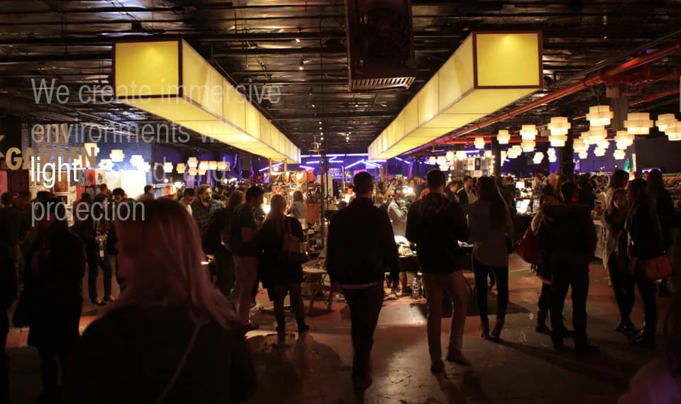 Crowd walking through an indoor market-like space lit with warm lantern lights and hanging paper lamps at a Nuit Blanche New York event.