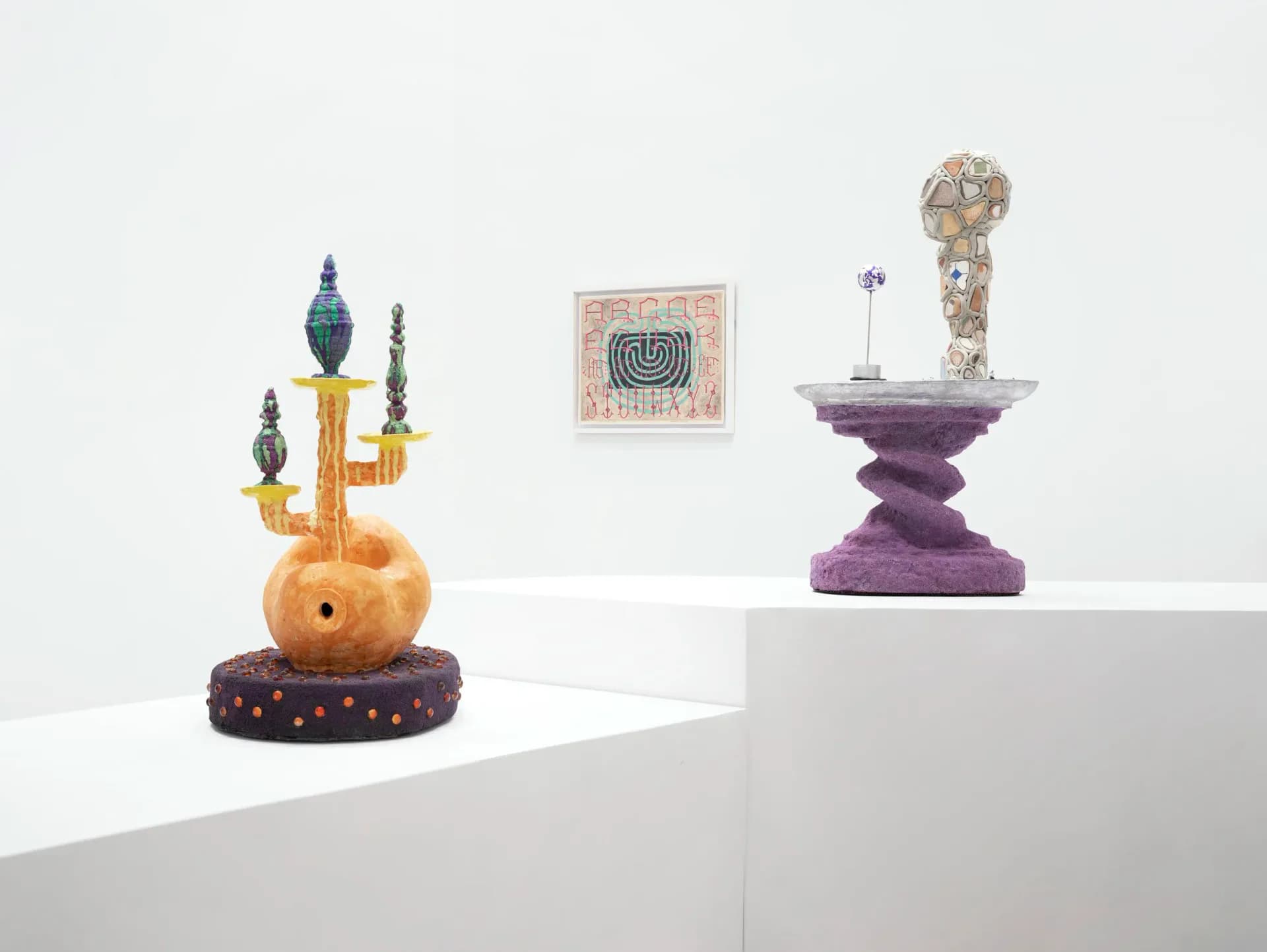 Gallery display at Nunu Fine Art with two sculptural objects on white pedestals: orange sphere with dark spires and purple geometric form, plus small framed artwork.