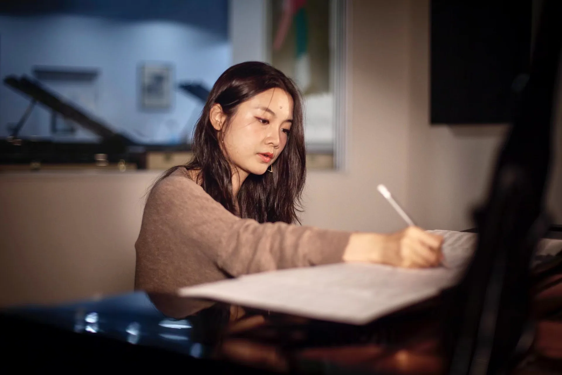 Person with dark hair seated at a desk, holding a brush or stylus, focused on work on a tablet or drawing surface in warm indoor lighting.