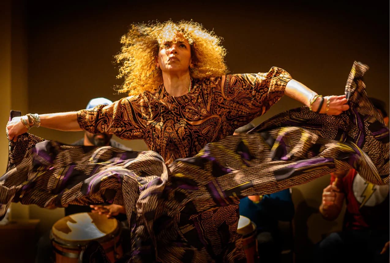 Performance or installation artwork featuring a figure with voluminous curly hair in a patterned bronze outfit, arms outstretched amid draped fabrics and dramatic stage lighting.