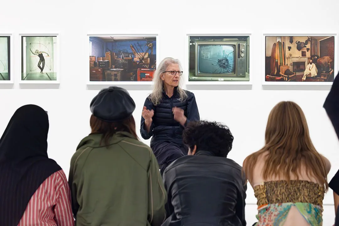 Gallery presentation with woman in dark vest speaking to a group of visitors standing before framed artworks on white wall.