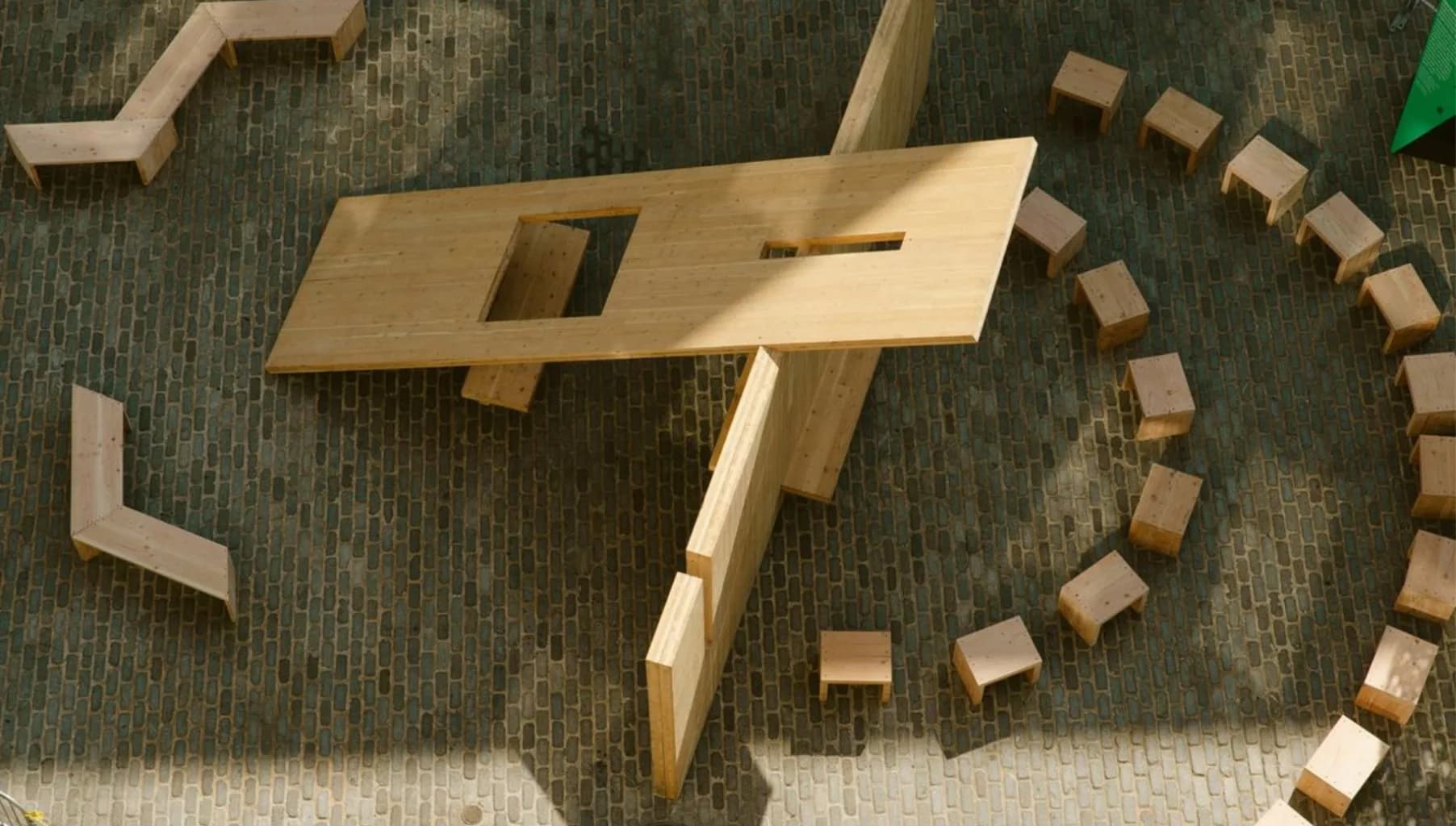 Wooden sculptural form with geometric cutouts and curves, surrounded by small cubic wooden blocks arranged in curved pattern on carpeted floor.