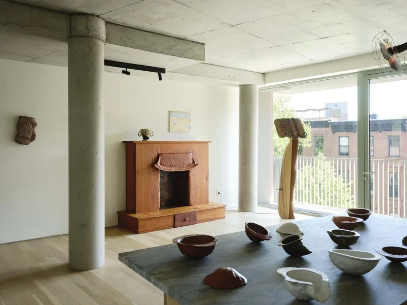 Gallery interior with concrete column, wooden sculptural fireplace, ceramic vessels on gray table, and large windows overlooking cityscape.