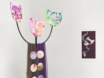 Mixed-media sculpture: wooden vase with painted circular elements holding three pink and green fabric flowers with black stems; small framed drawing on wall.
