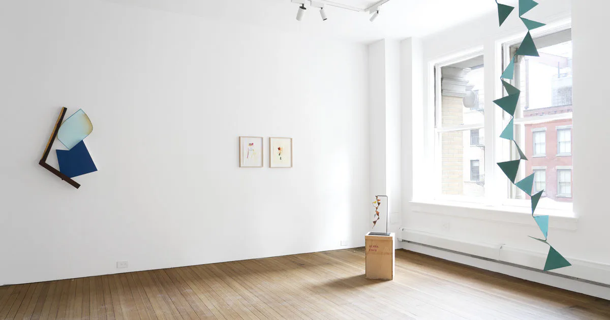 Bright gallery interior with white walls, wooden floor, geometric paper decorations hanging from window, and small sculptural objects on display.
