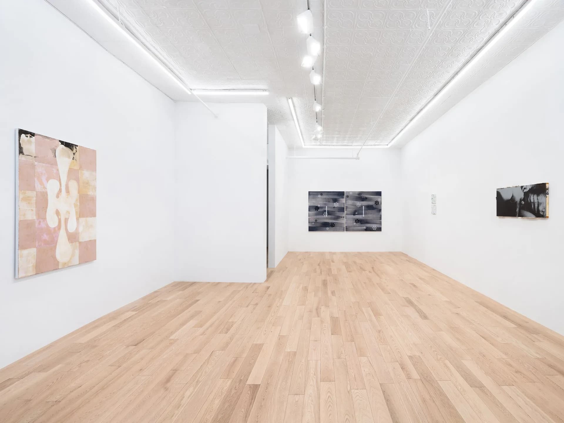 Clean white gallery interior with light wood flooring, three artworks on walls including a pink abstract piece on left, and black and white photographs on opposite wall.