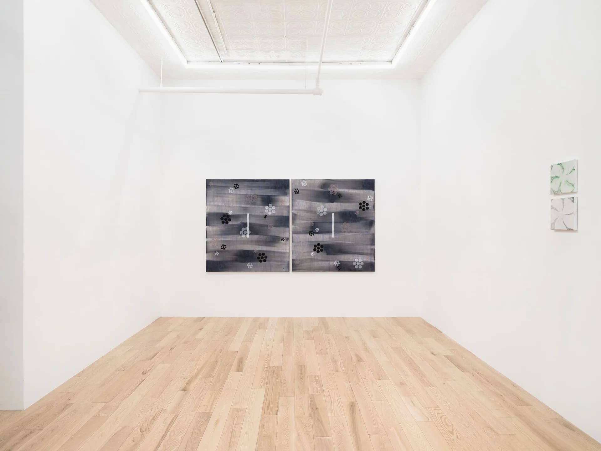 White gallery space with light wood flooring, black and white diptych photograph on wall, small artwork visible on right wall.