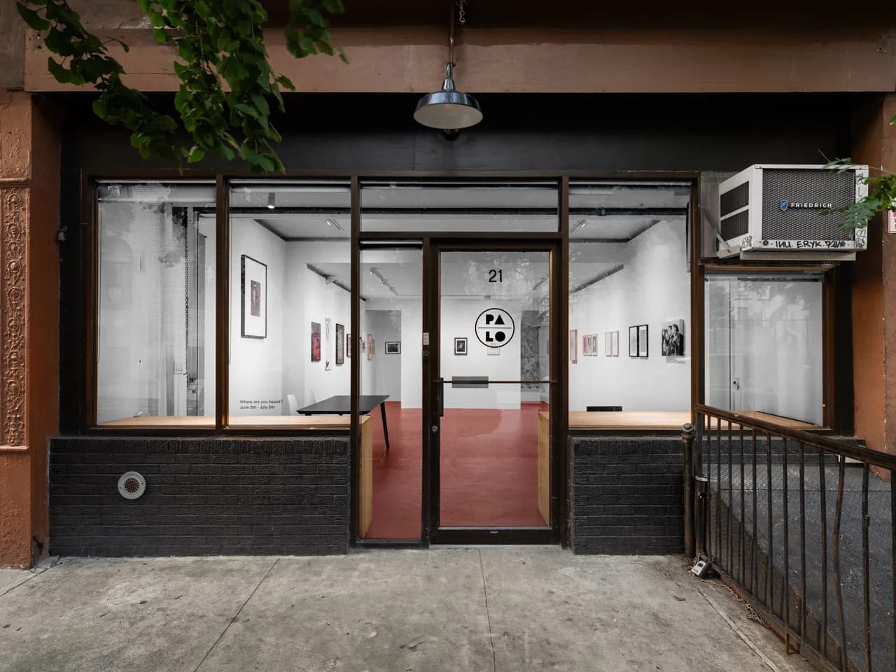 Storefront entrance of Palo Gallery with glass windows revealing white-walled interior hung with framed artworks and a red floor