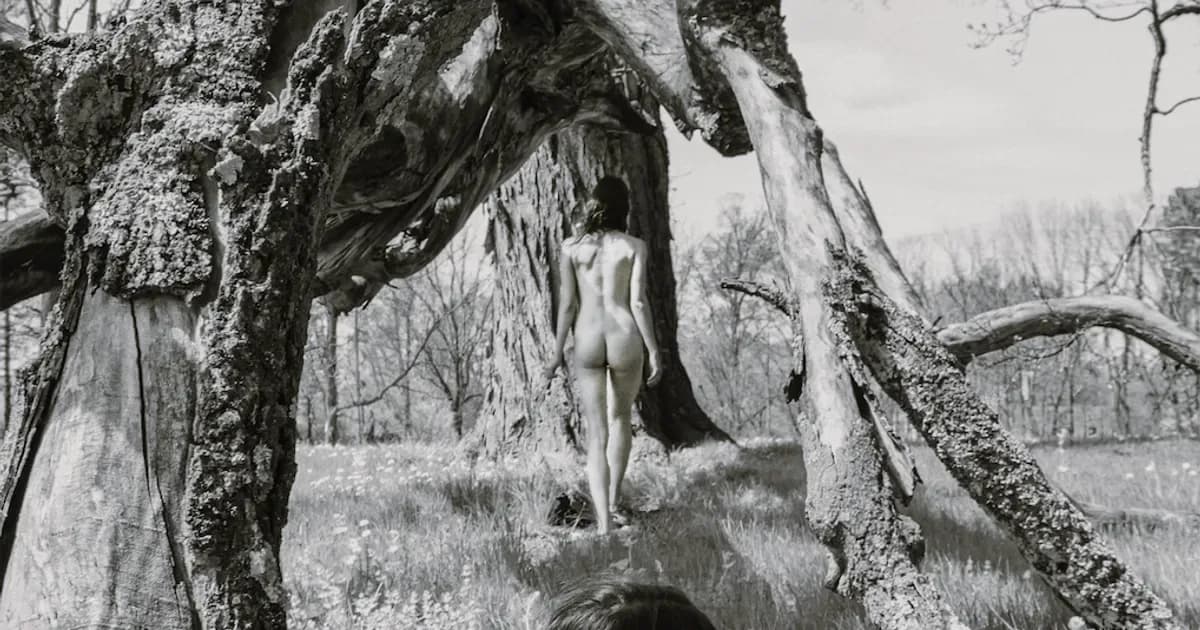 Black and white photograph of a nude figure standing beneath gnarled tree branches in a sparse woodland landscape.