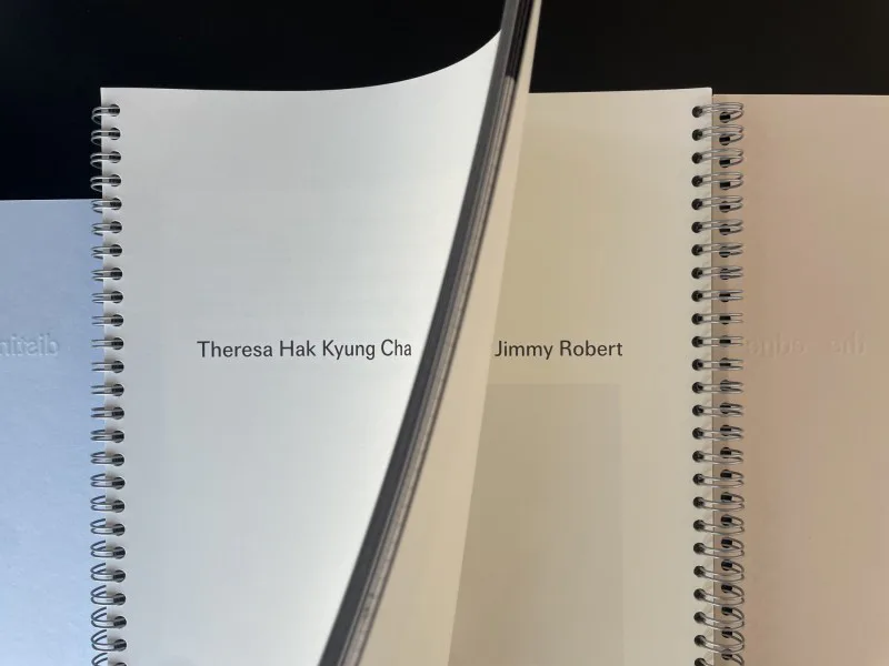 Two open spiral notebooks displaying artist names 'Theresa Hak Kyung Cha' and 'Jimmy Robert' on facing pages.