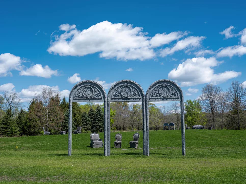 Three arched metal structures with ornamental tops installed in a grassy outdoor field against a blue sky with white clouds.