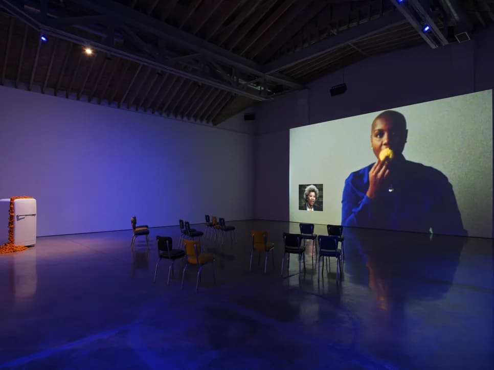 Gallery interior with purple-tinted lighting, projection screen showing a man's face, scattered colorful chairs, and minimalist furnishings.