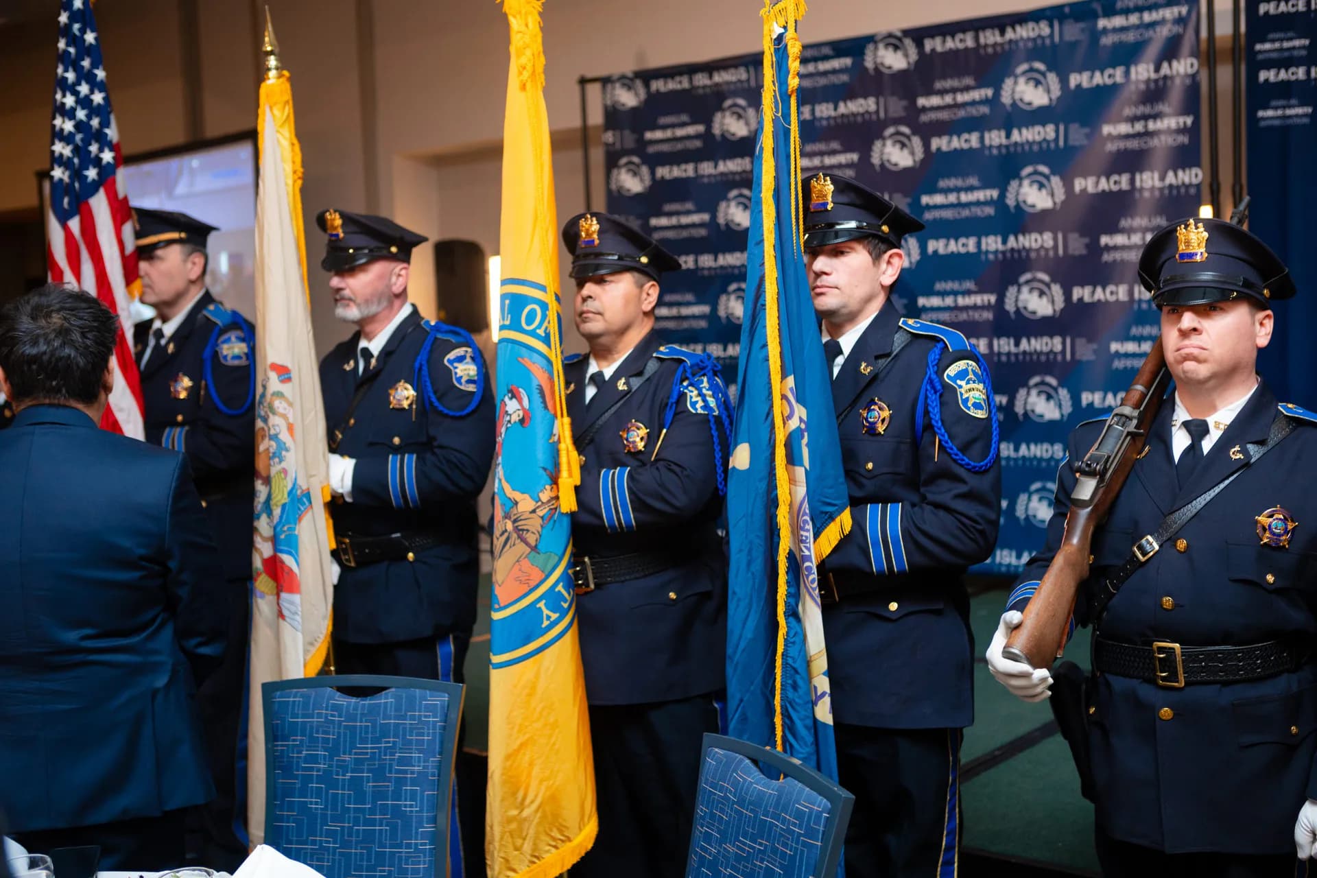 Military personnel in dress uniforms holding flags at Peace Islands Institute event, with American flag and institutional backdrop visible.