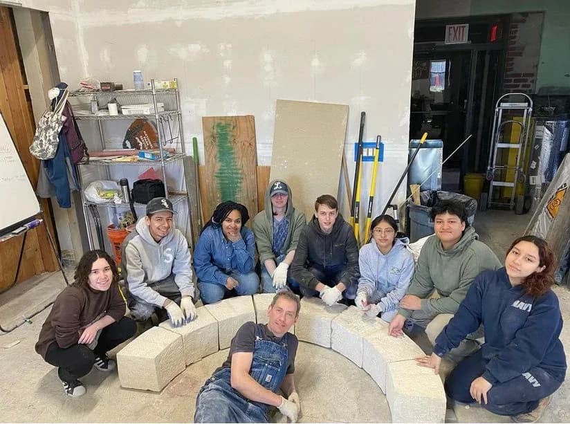 Group of students and instructors posing around a curved stone bench they built inside the Pellettieri Stone Carvers' Academy studio