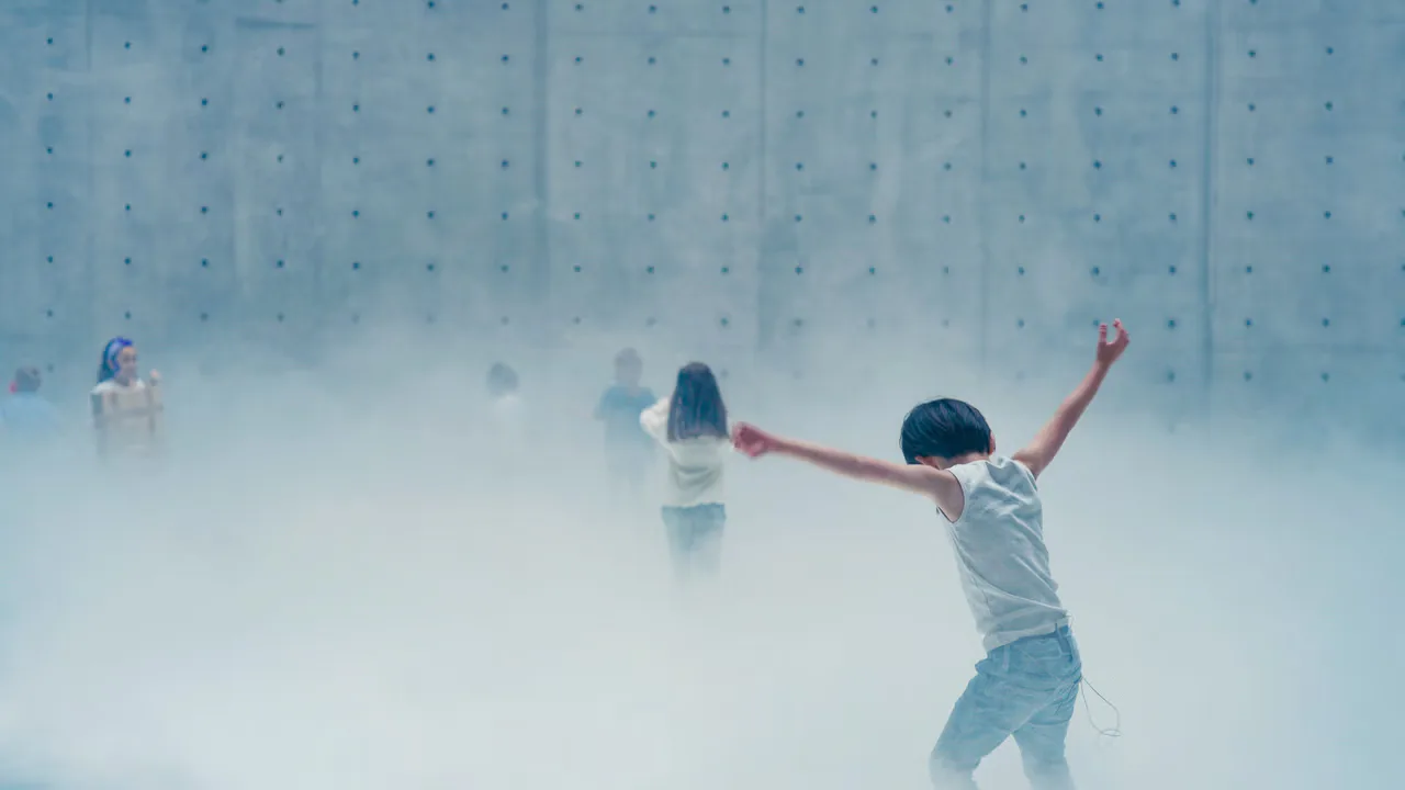 Children in white clothing dance and gesture with raised arms in a misty, fog-filled indoor space with dotted wall pattern.
