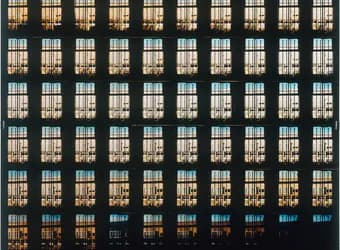 Large-scale abstract photograph or installation featuring a grid of window-like forms with warm golden and cool blue tones alternating across dark architectural framework.