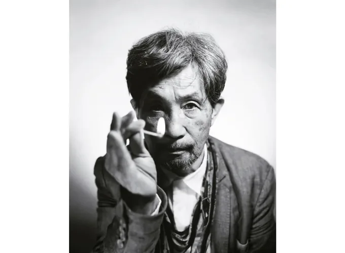 Black-and-white portrait photograph of an Asian man with short dark hair, wearing a collared jacket, gesturing with his hand toward the camera.