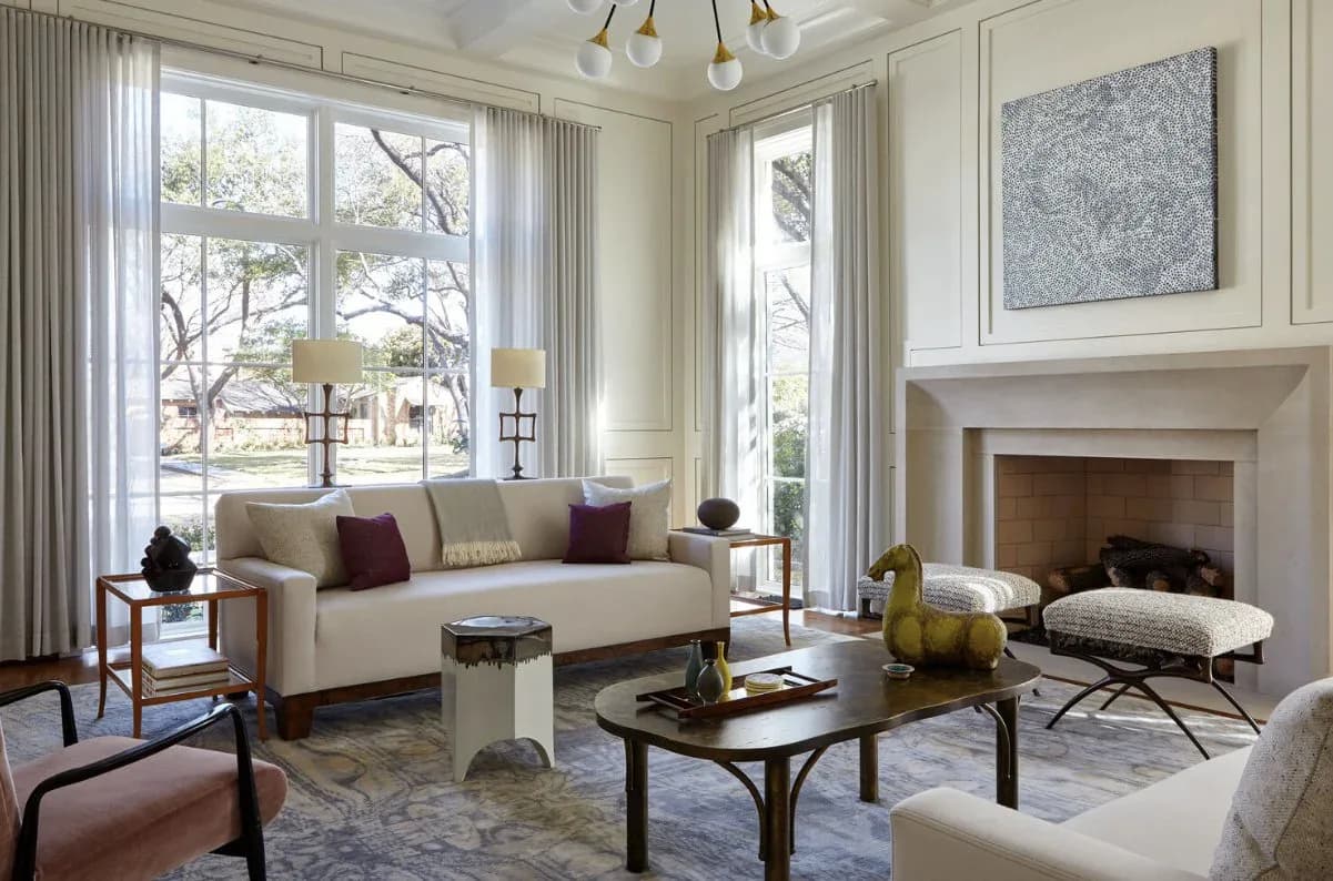 Elegant living room with cream sofa, fireplace, large windows with sheer curtains, dark coffee table, and blue-toned abstract artwork above mantel.