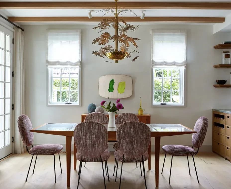 Bright dining room with wooden table, four upholstered chairs, exposed beam ceiling, large windows, and minimalist wall art.