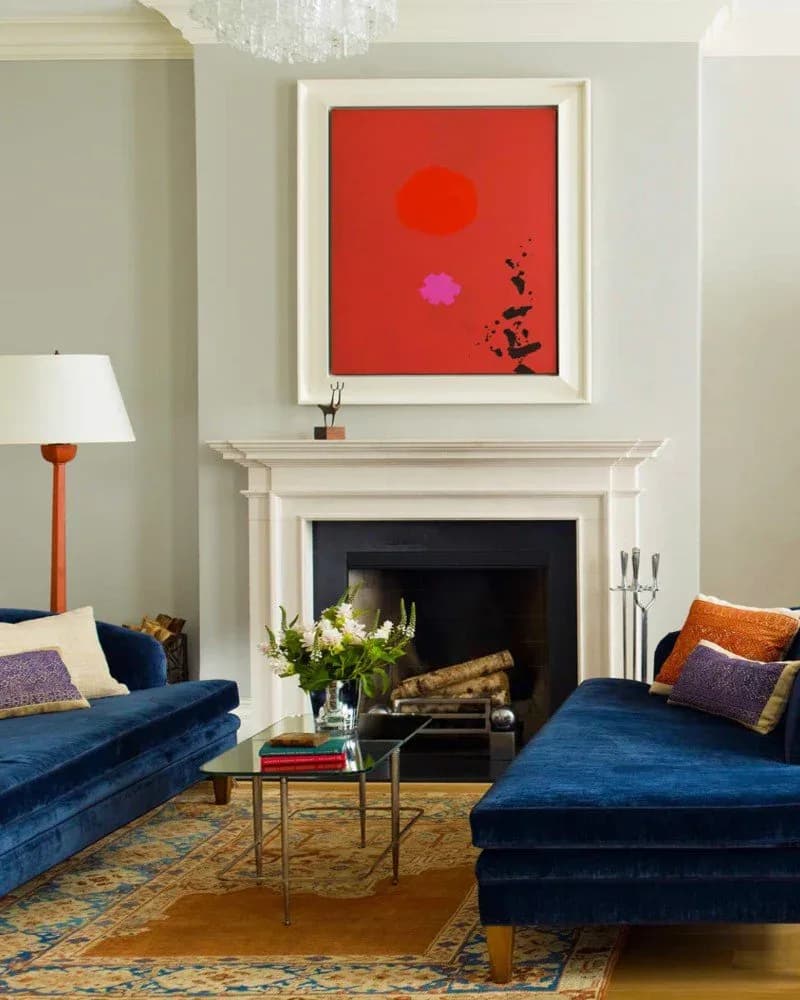 Living room with pair of blue velvet chaise lounges, white fireplace with red and pink abstract artwork, and patterned rug.