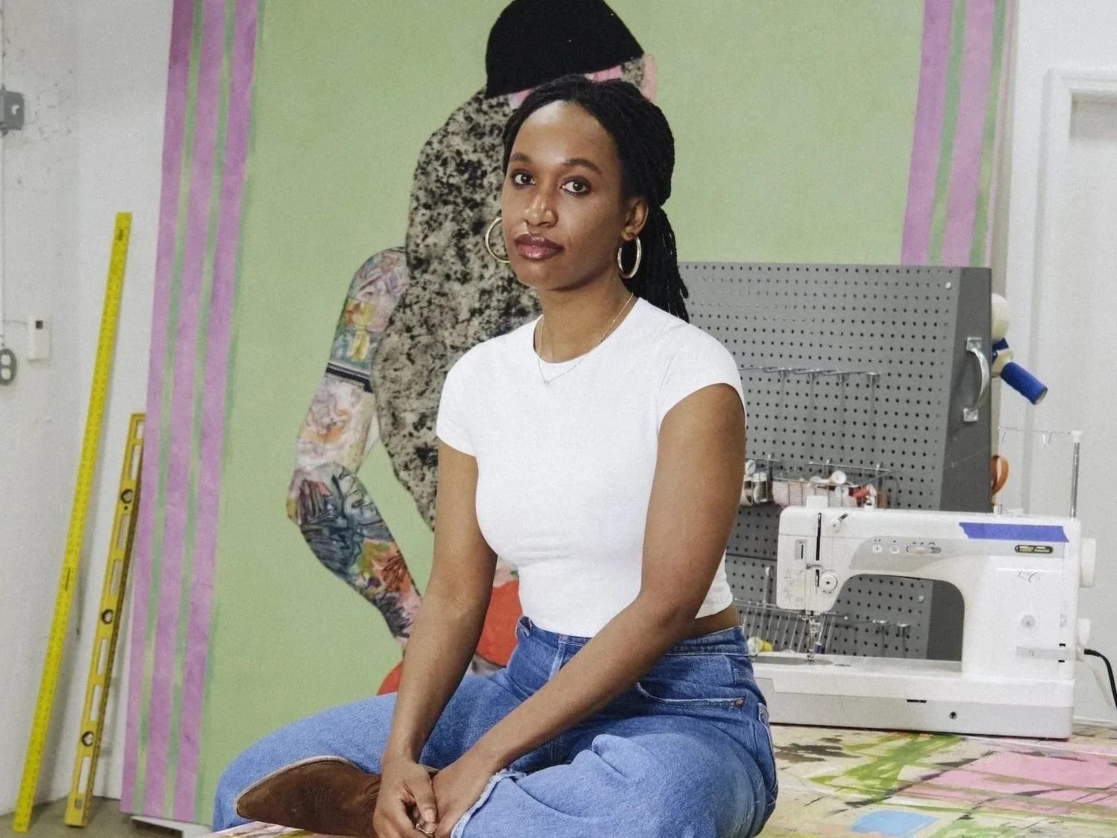 Portrait of a woman in white sleeveless shirt and black beanie sitting in a studio space with sewing machine and colorful fabric visible.