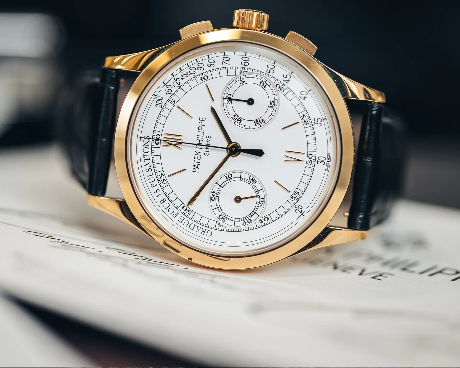 Gold chronograph wristwatch with white dial, Roman numerals, and black leather strap, displayed on white paper.