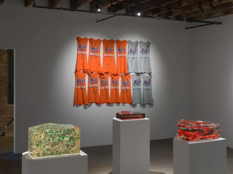 Installation with orange and gray fabric works mounted on white wall above three pedestals displaying colorful textile or mixed-media pieces.