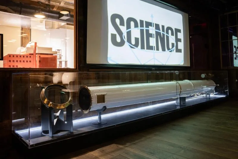 Gallery interior with illuminated display case, word 'SCIENCE' projected on wall, architectural model visible, dark wood flooring.