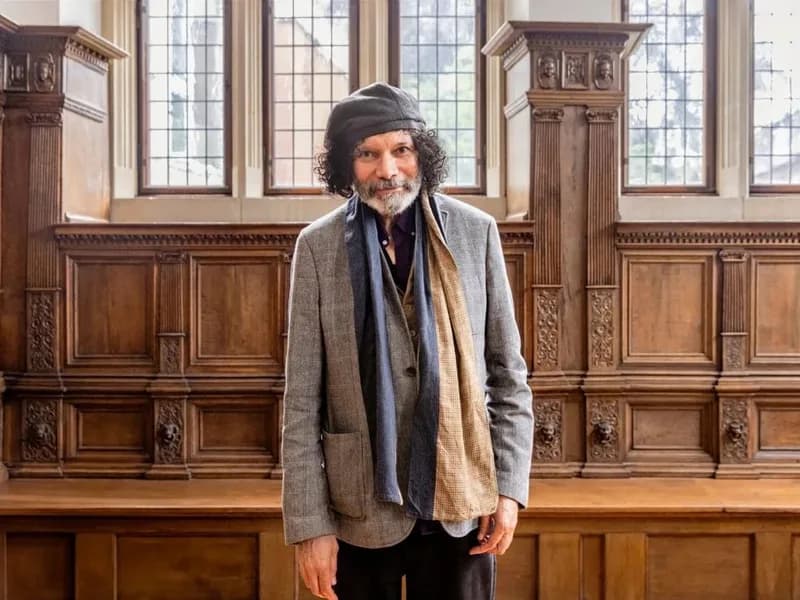 Man with dark wavy hair and beard wearing a gray blazer and dark scarf, standing in a wood-paneled gallery space with tall windows.