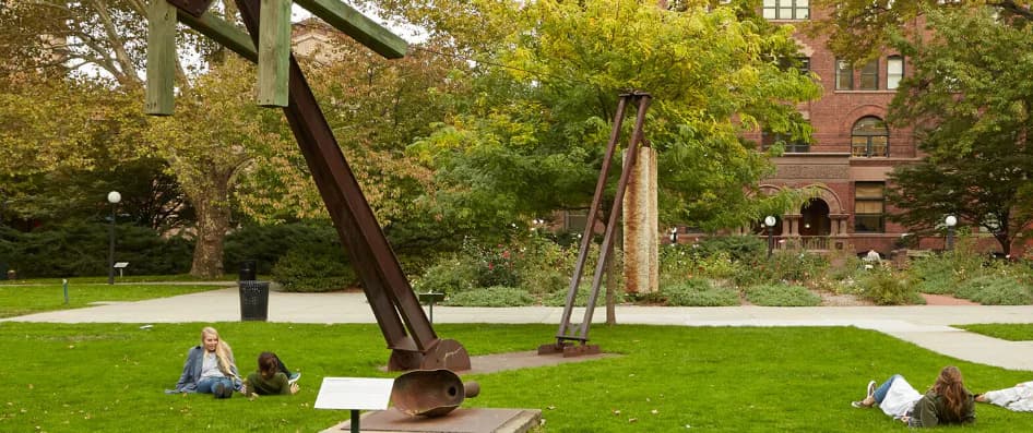 Outdoor lawn at Pratt Institute Sculpture Park with abstract metal sculptures and brick academic building in background; autumn foliage visible.
