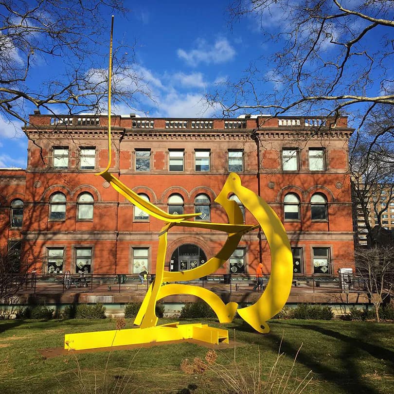 Large-scale yellow abstract sculpture with curved forms installed on lawn in front of red brick building; metal construction.