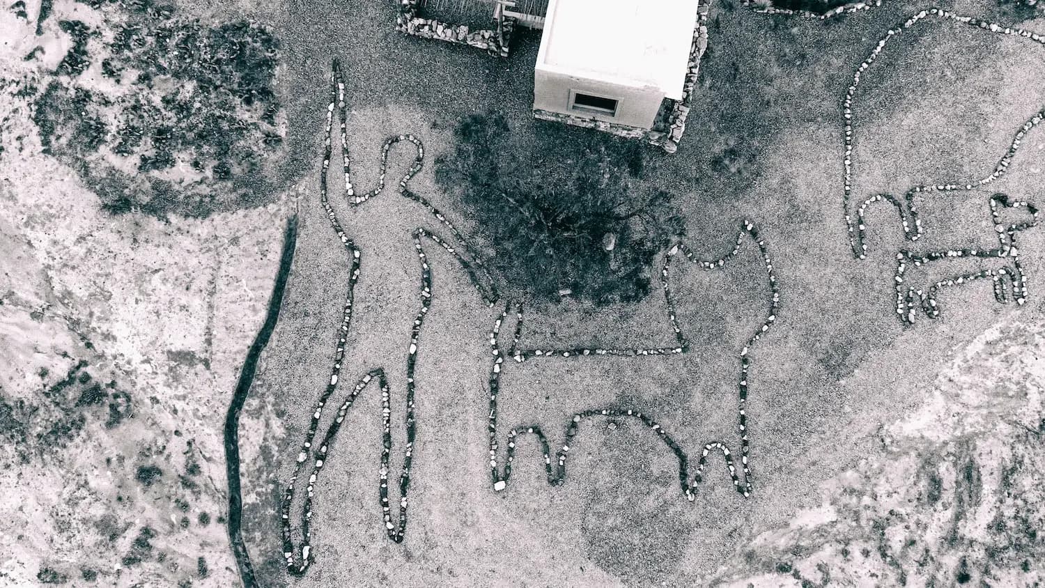 Aerial photograph of land with geometric patterns marked by stones or vegetation, featuring a white structure and winding path.