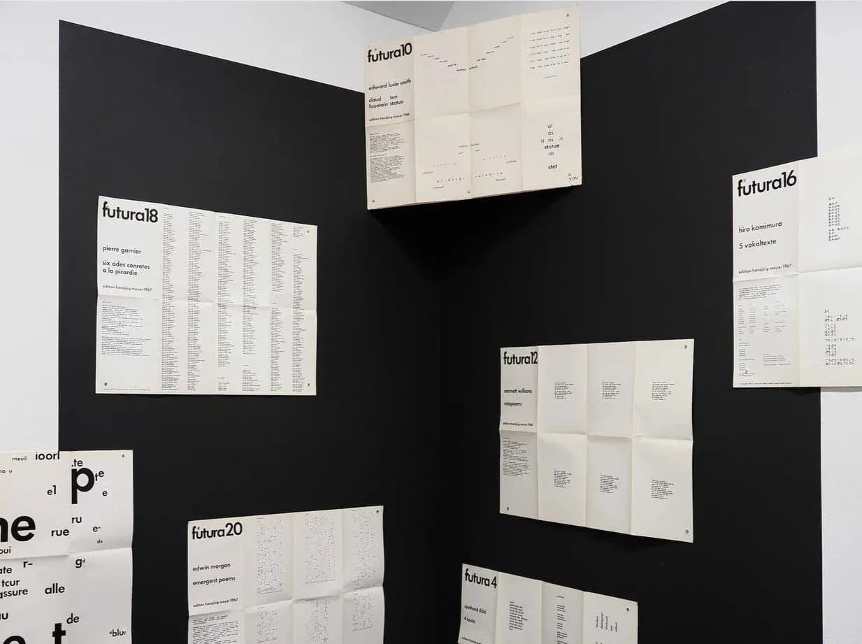 Interior gallery view with black walls displaying multiple white printed sheets and documents labeled "futura18," "futura20," and "futura21," minimalist exhibition setup.