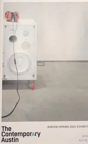 Installation artwork featuring a white speaker-like panel with headphones and audio components mounted on a gray wall, from The Contemporary Austin Winter/Spring 2015 exhibition.