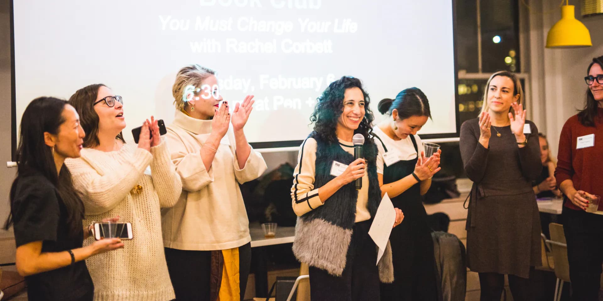 Group of people applauding indoors at an event with a projection screen visible in background.