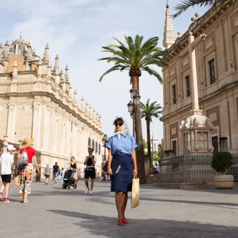 Outdoor street scene of a grand European plaza with ornate stone buildings, palm trees, and pedestrians walking.