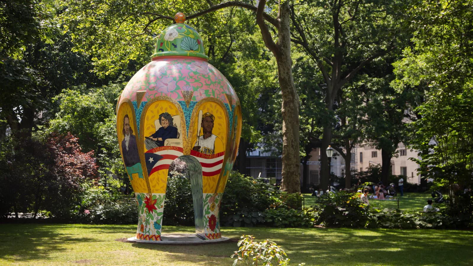 Large colorful spherical sculptural installation with yellow, orange, and blue arched openings, positioned in a lush green park setting with trees.