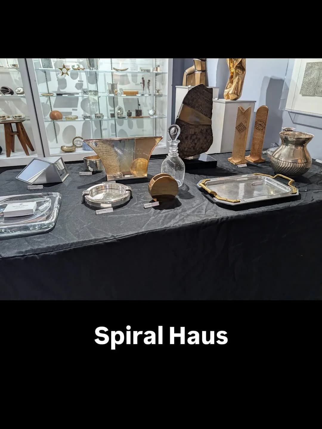 Gallery display table at Spiral Haus showing decorative objects including brass vessels, sculptural forms, and tableware on dark marble surface with glass cases and artwork visible in background.