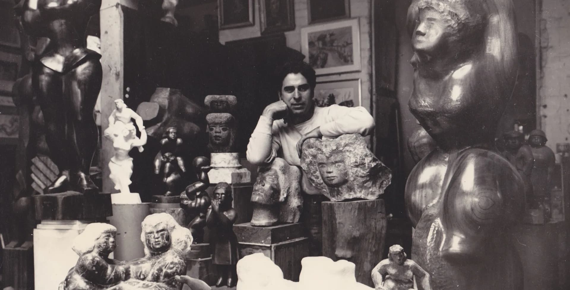 Black and white photograph of a studio interior with a seated artist surrounded by sculptural works, busts, and figurative sculptures on shelves and tables.
