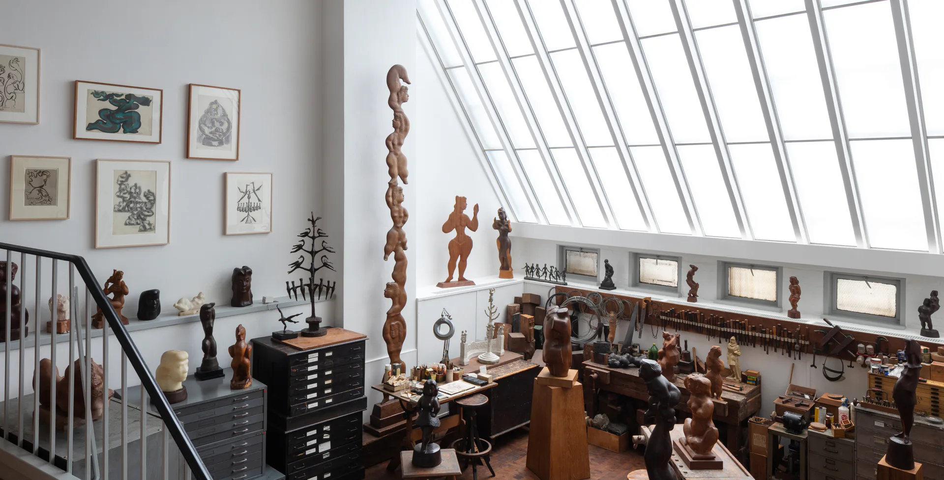Modern gallery interior with sloped skylight, wooden sculptural works displayed on pedestals and shelves, framed botanical prints on white walls.