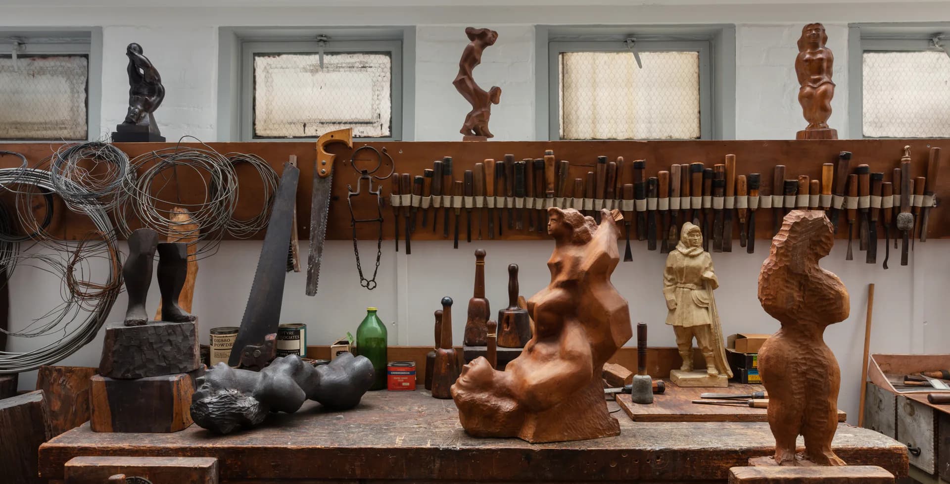 Studio worktable with wooden carved figures and sculptures in progress, tools, and materials arranged against white walls with sculptural works mounted above.