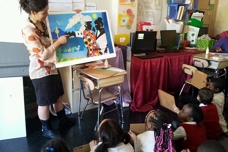 Interior photograph of a studio or classroom showing a woman holding an artwork while children sit on the floor, with shelves and art supplies visible in the background.