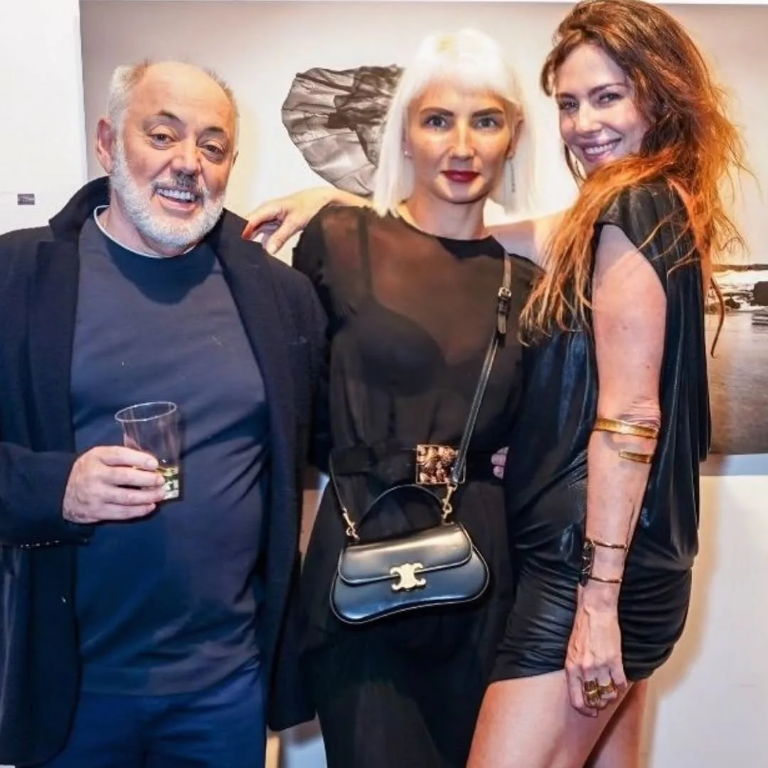 Three people at an indoor event: an older man in dark navy, a woman with platinum blonde hair in black, and a woman with reddish-brown hair in black leather.