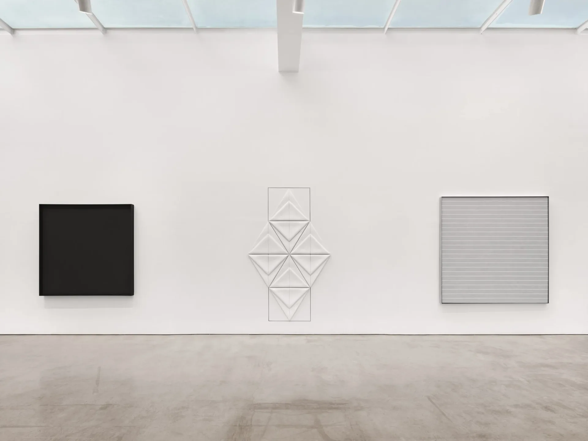 Gallery interior with three artworks on white wall: black square, geometric line drawing, and gridded square.