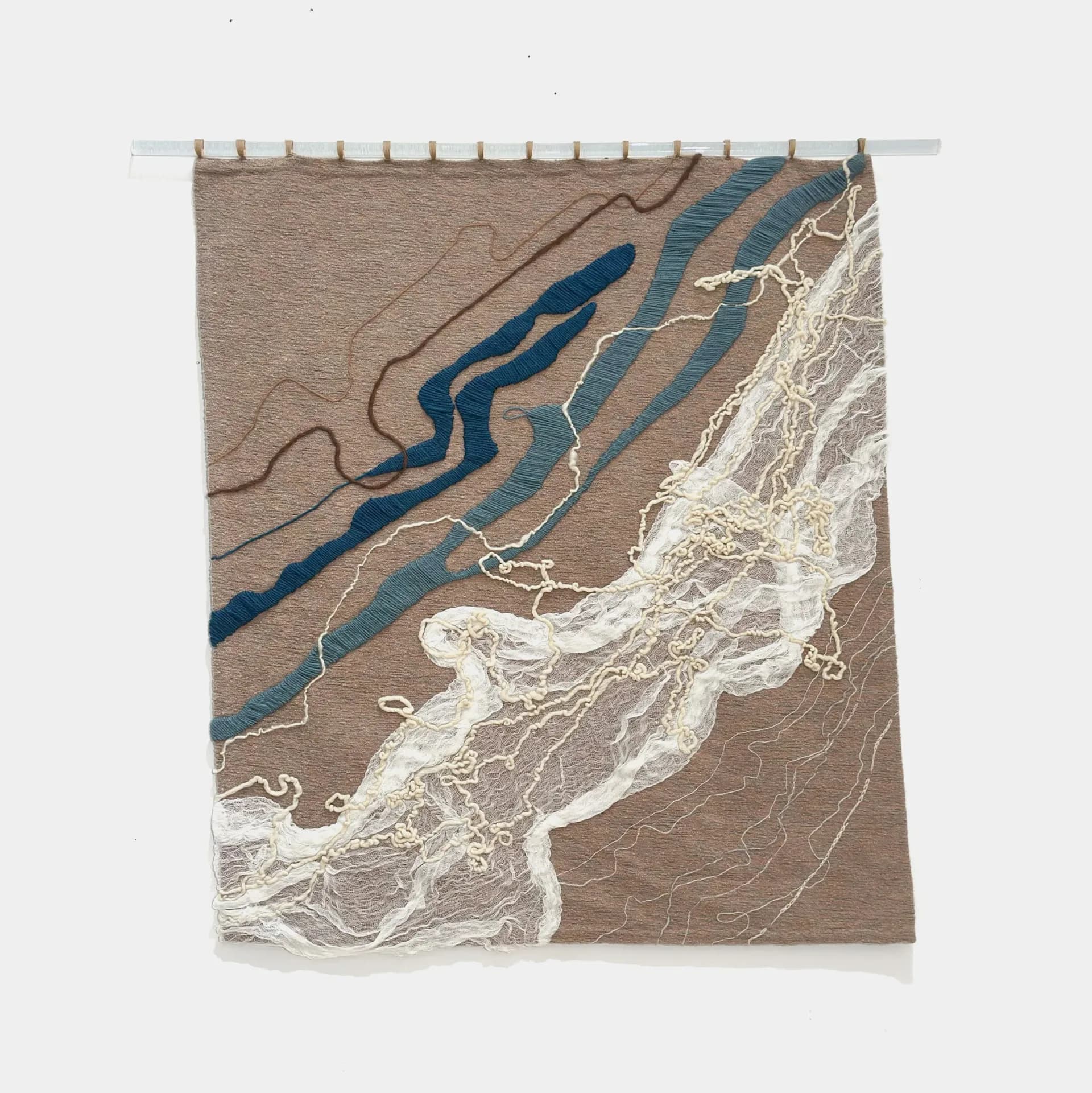 Textile wall hanging depicting abstract landscape with blue river forms, tan striped sections, and white lace-like mountain or wave imagery against beige ground.