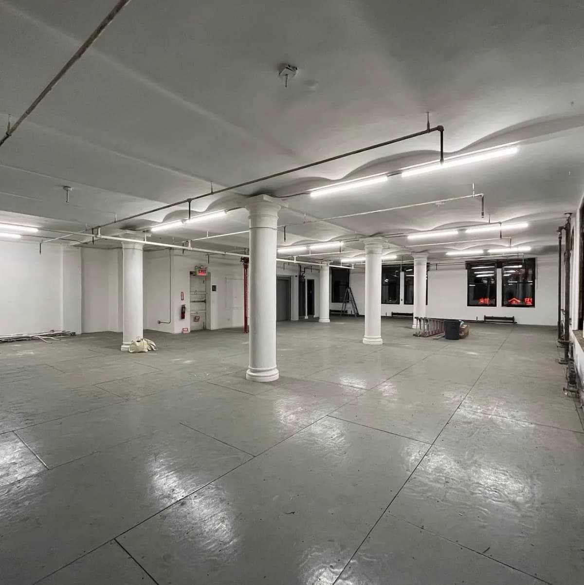 Empty gallery space with white columns, concrete floors, exposed ceiling pipes, and rows of fluorescent lighting.